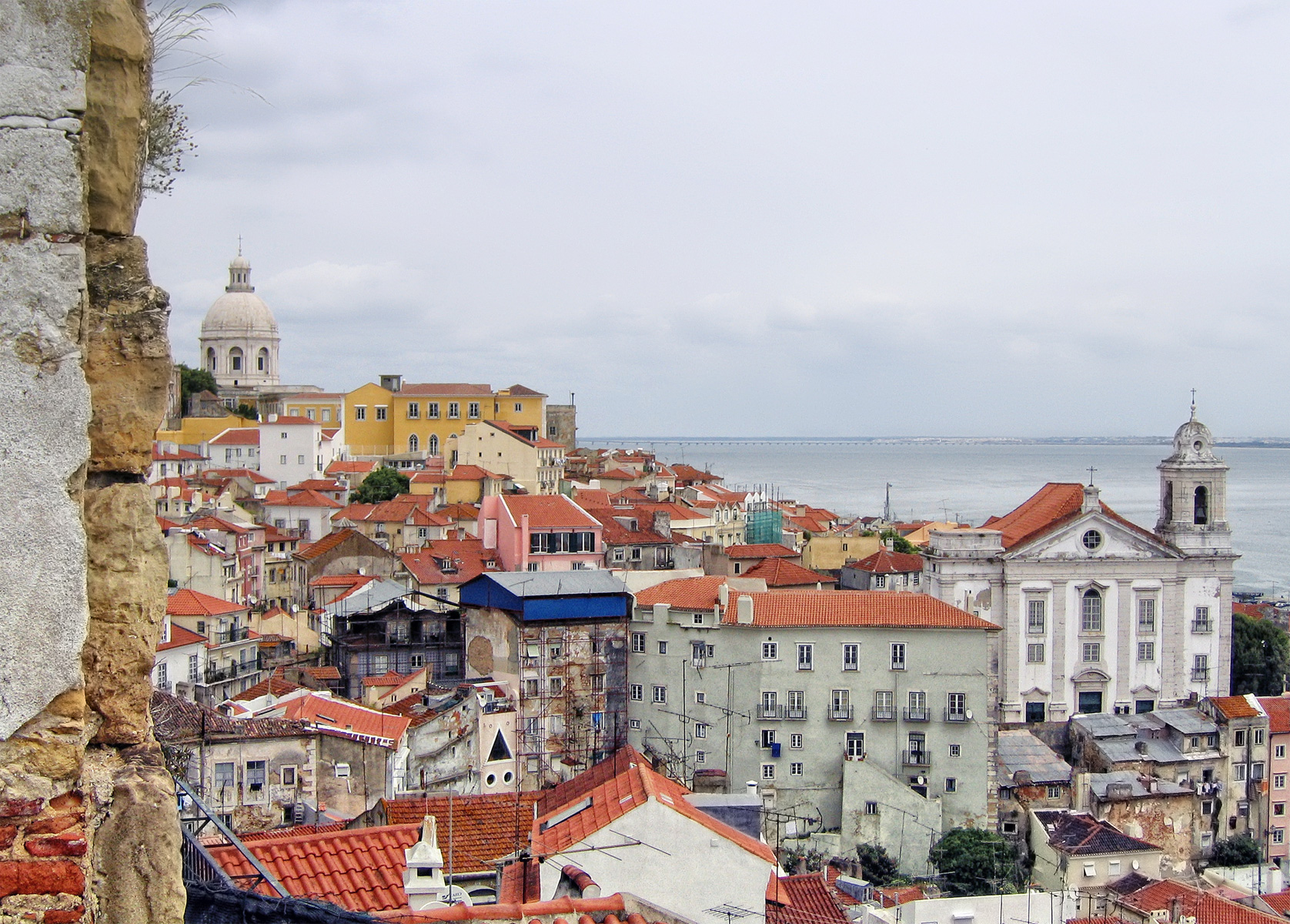 Vista panorâmica de Lisboa com telhados vermelhos, edifícios históricos e o rio Tejo ao fundo.